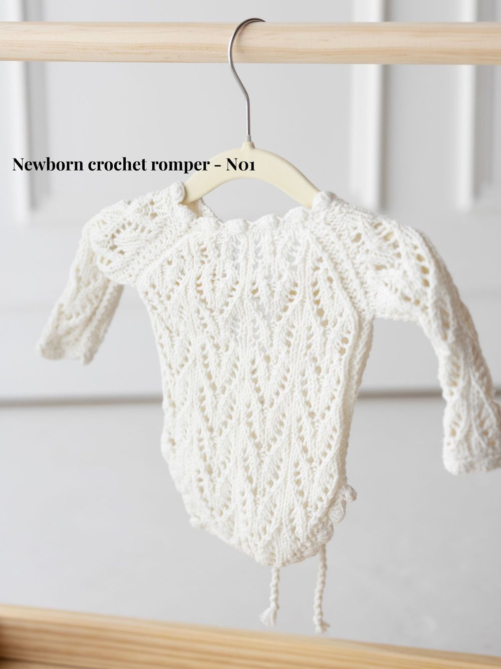 Delicate white crocheted newborn romper with intricate lace patterns displayed on wooden hangers against a light background.