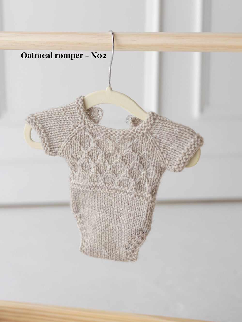 Handmade knit baby romper in oatmeal color displayed on wooden hangers showing different angles of the garment.