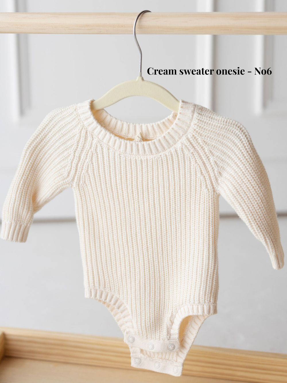 Cream colored knit baby sweater onesie displayed on wooden hanger against white wall background.