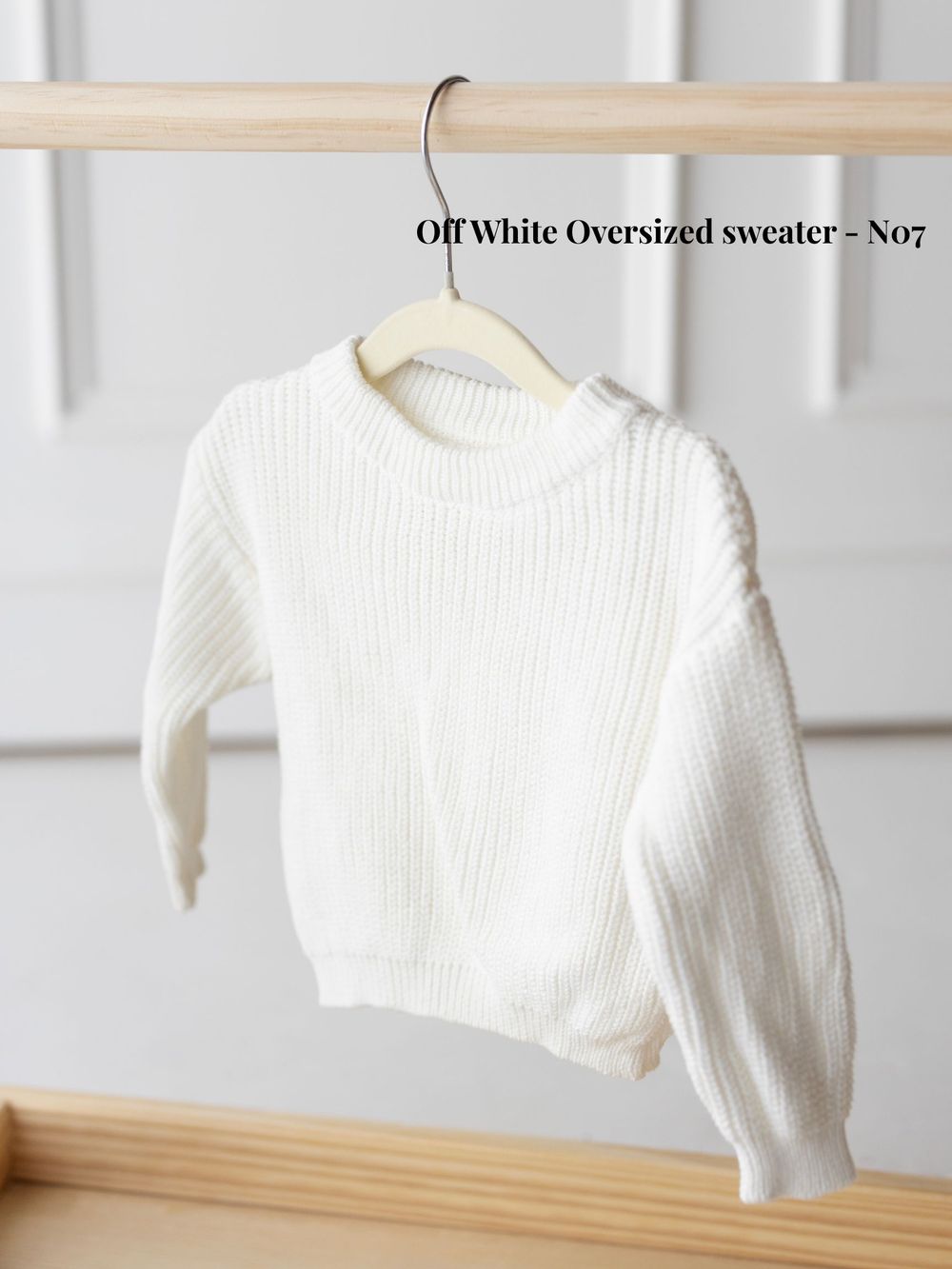 White oversized knit sweater hanging on wooden clothing rack against light background.
