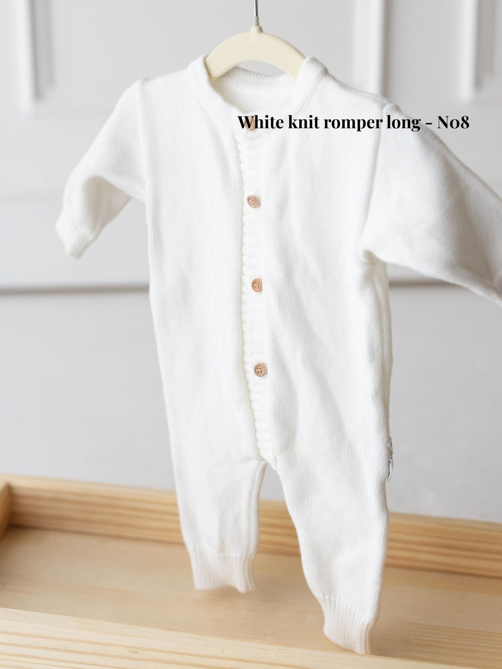 White knit baby romper with buttons displayed hanging on wooden hangers against a light background.