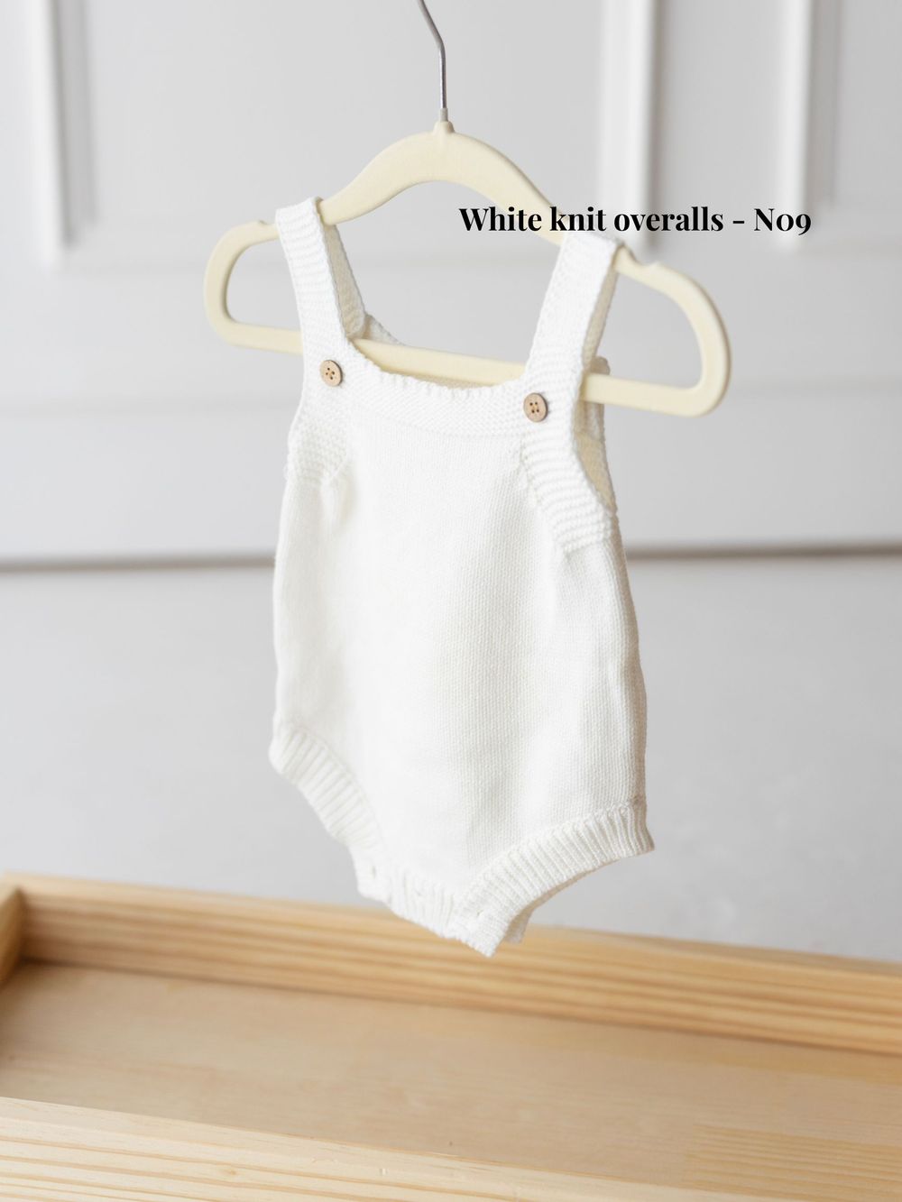 White knitted baby romper overalls hanging on display against a light background.