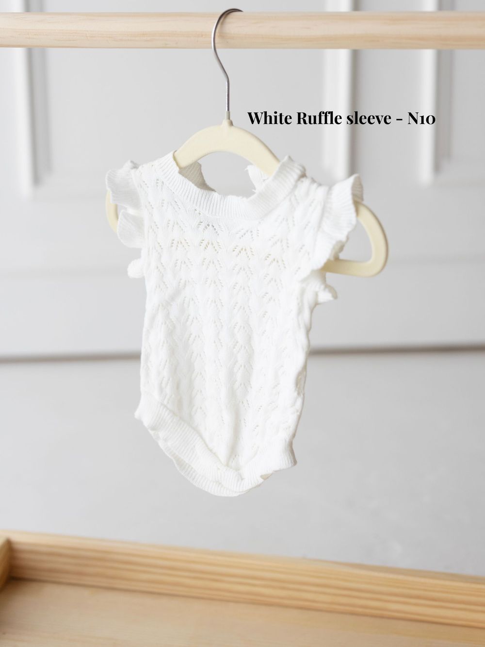 White knitted baby bodysuit with ruffle sleeves hanging on wooden hanger against light gray wall.
