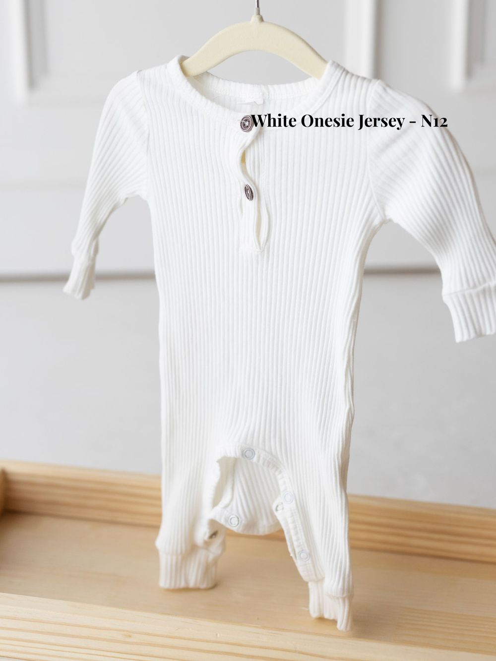 White ribbed jersey onesie for infants hanging on wooden display hanger against neutral wall.