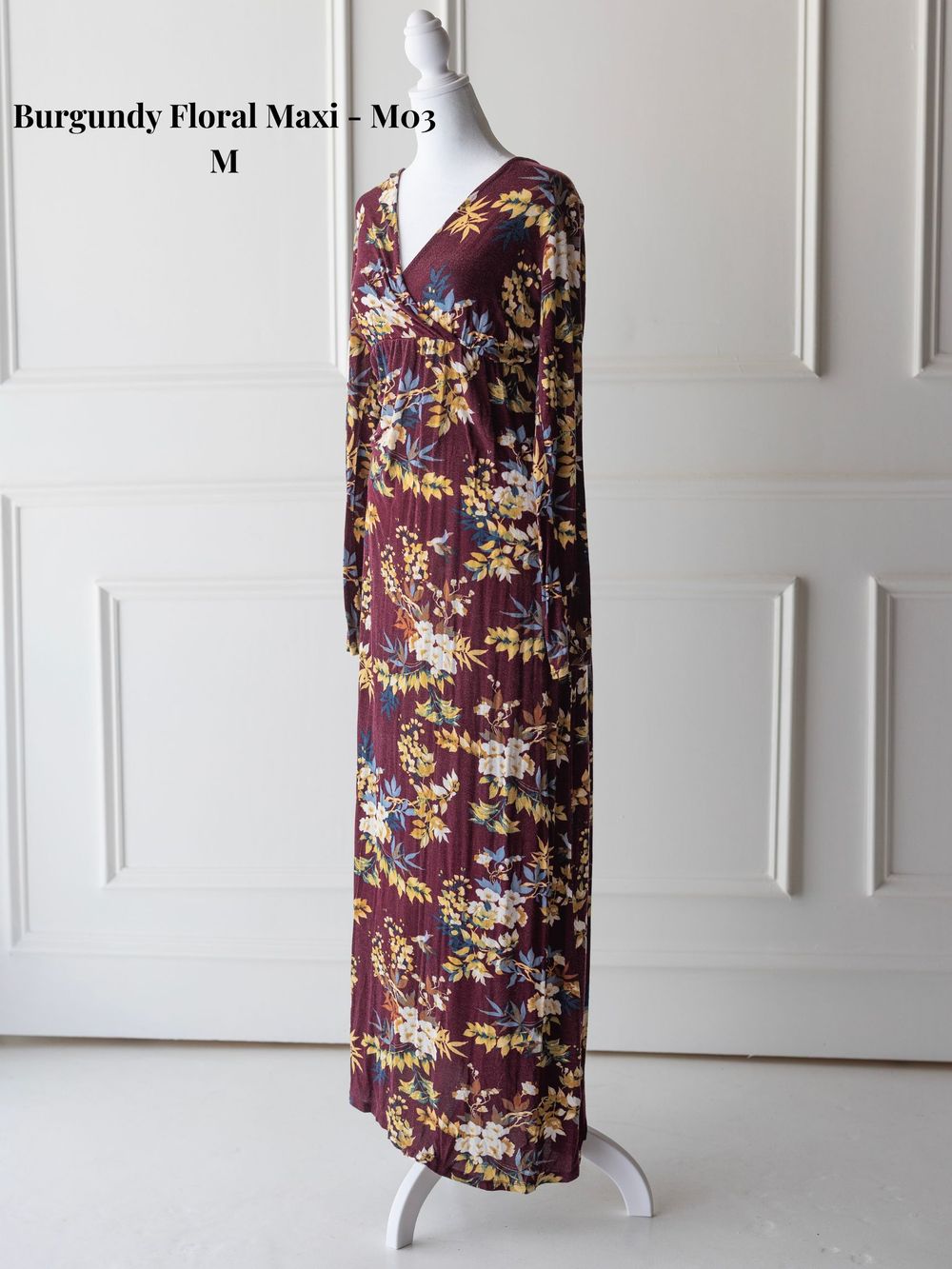Burgundy floral maxi dress with long sleeves and empire waist shown on mannequin against white paneled wall.