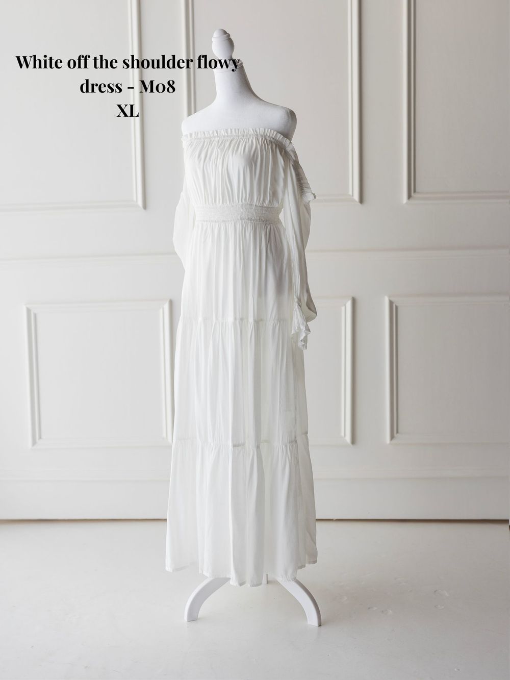 White off-shoulder flowy maxi dress displayed on mannequin against white paneled wall in multiple views.