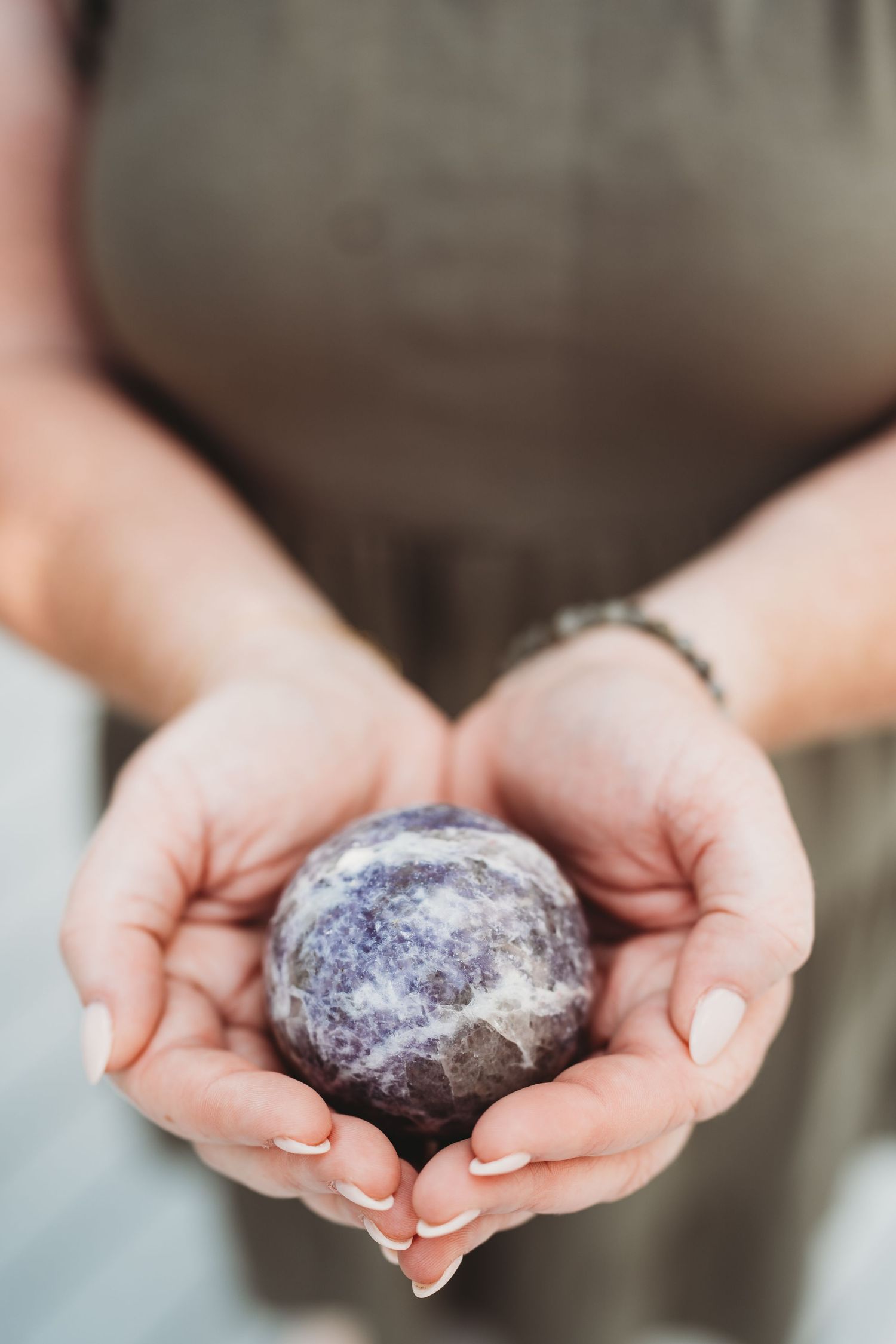 Hands gently holding a polished amethyst crystal sphere against a soft blurred background.