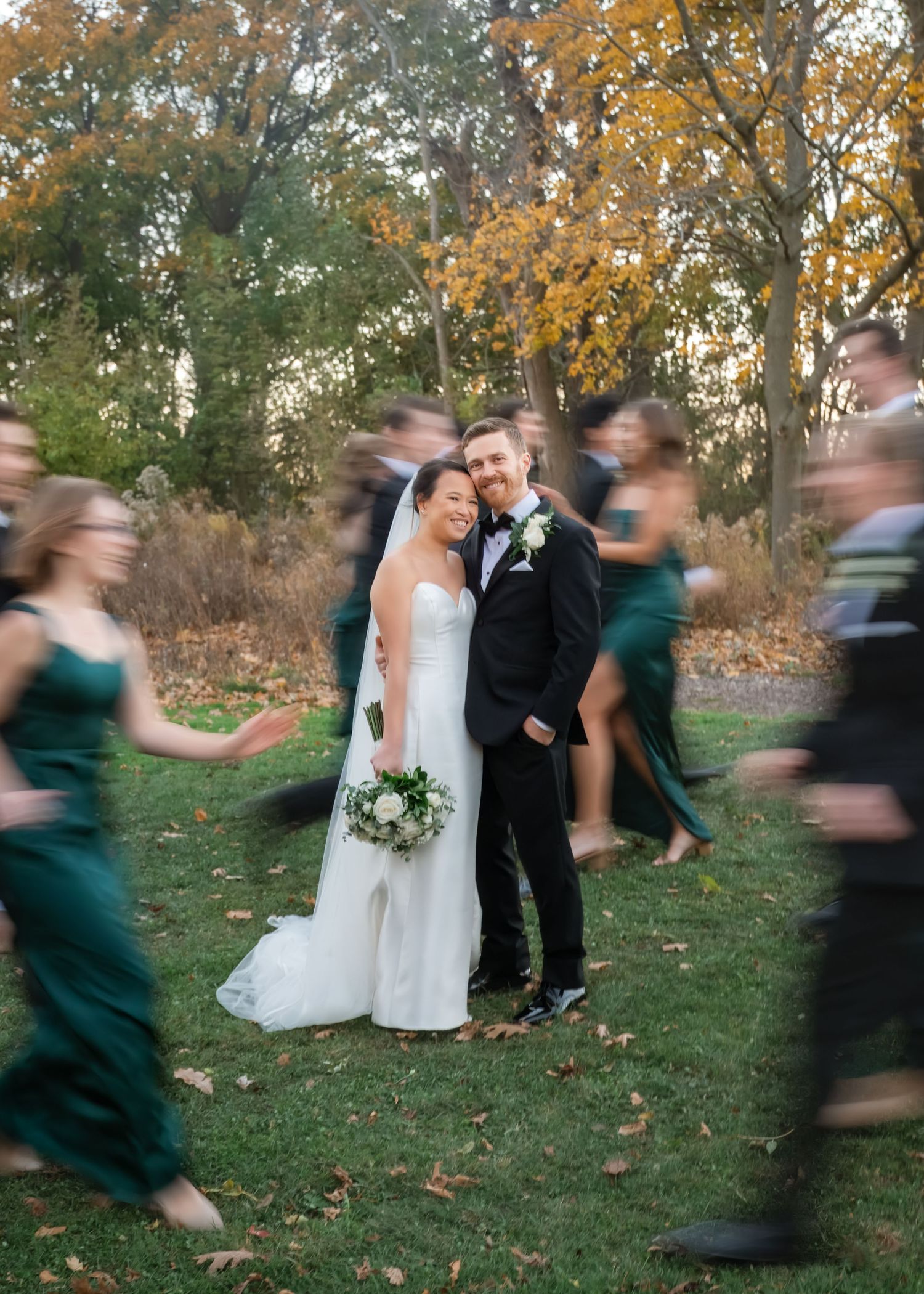 Creative wedding photo showing couple standing still while bridesmaids in emerald dresses create motion blur around them.