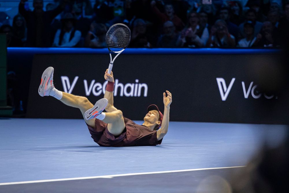 Tennis player falls backwards during intense match on indoor court with VeChain advertising visible.
