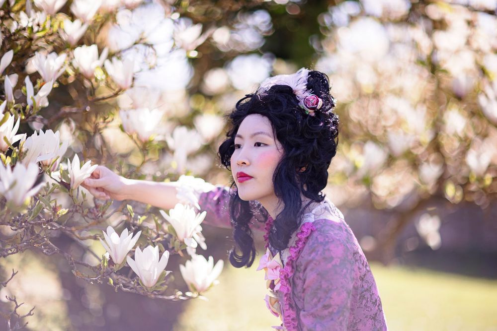 Woman in traditional Asian costume with floral hair accessories touches white magnolia blossoms in a sunlit garden.