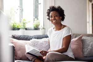 A person relaxes on a comfortable grey couch while reading a book near a bright window.