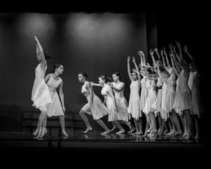 Black and white photo of dancers in white outfits performing in a line on a dark stage.