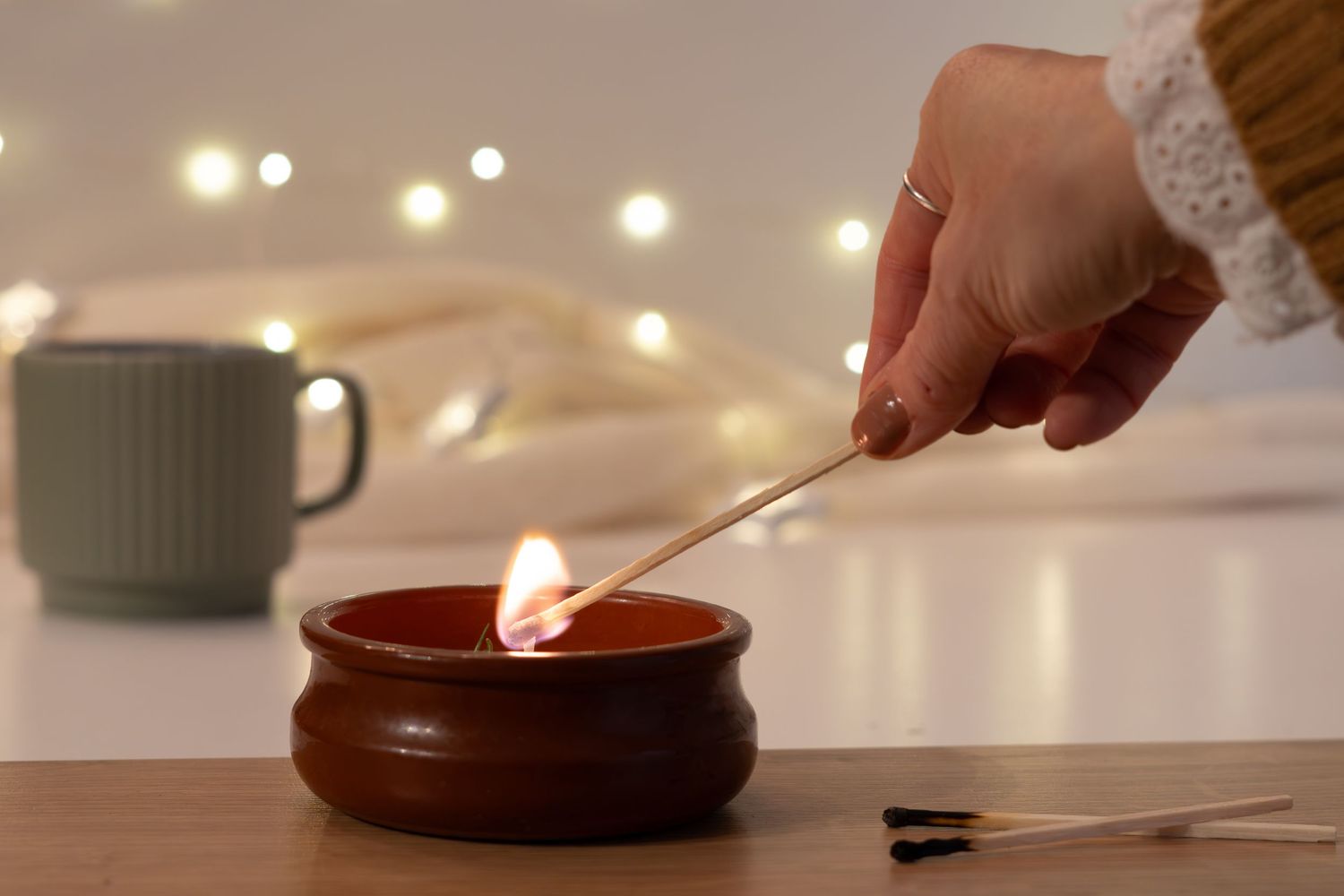 Still-life photograph of a glass jar candle being lit, arranged on a wooden table and photographed in natural window light