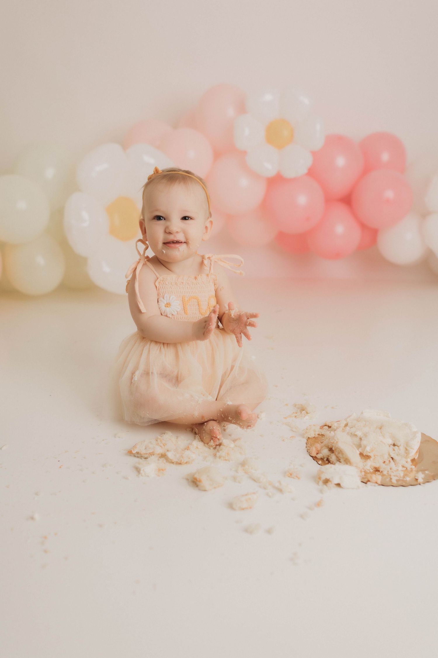 A baby in a pink dress sits on a light backdrop with pink and white balloons while playing with cake during a smash cake session.