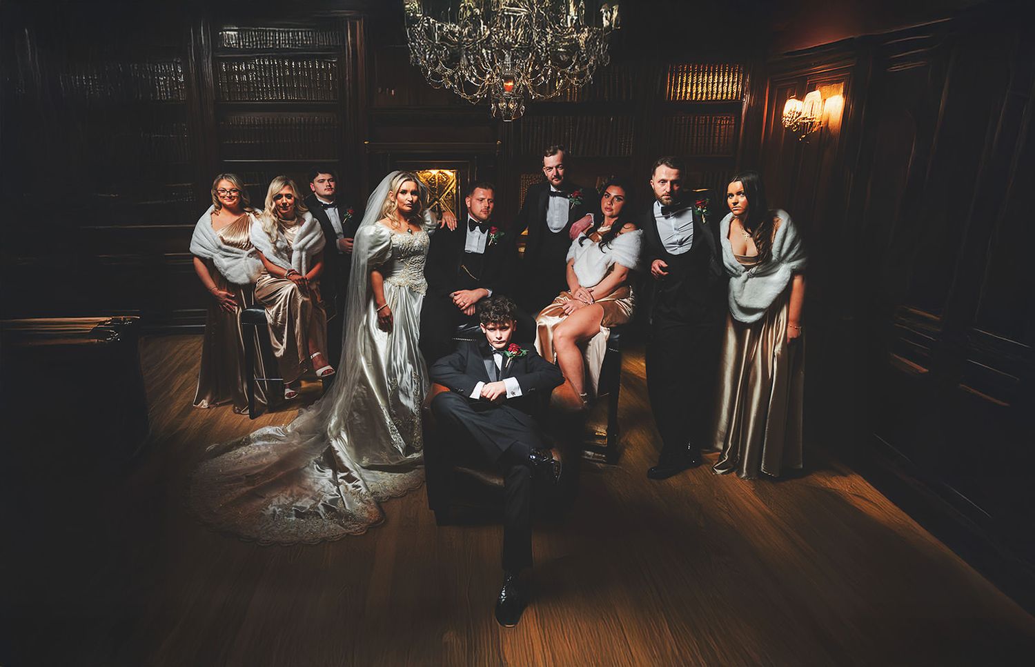 A wedding party poses together in a dimly lit room with elegant chandeliers and dark wood paneling.