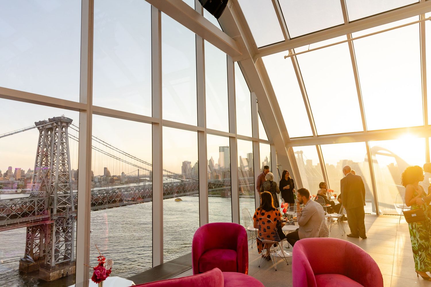 Panoramic sunset view through curved glass windows of a waterfront venue with bridge and skyline in background.