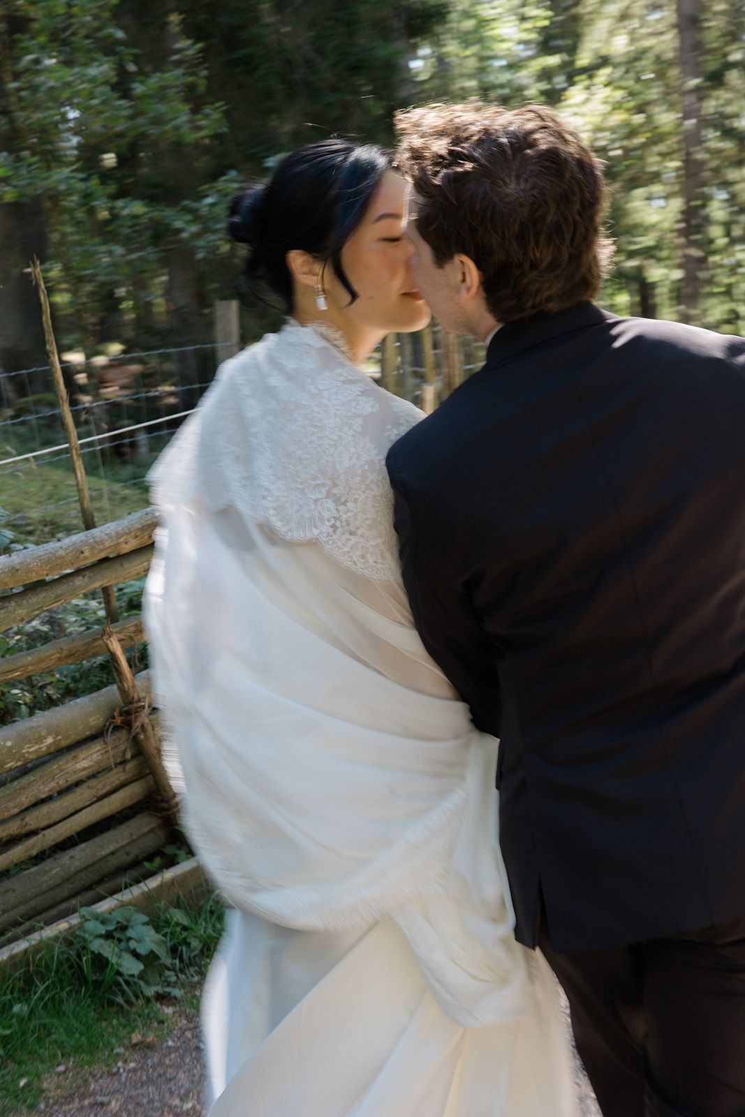 A romantic wedding moment captured outdoors with a bride in white dress and groom in black suit embracing near a rustic fence.