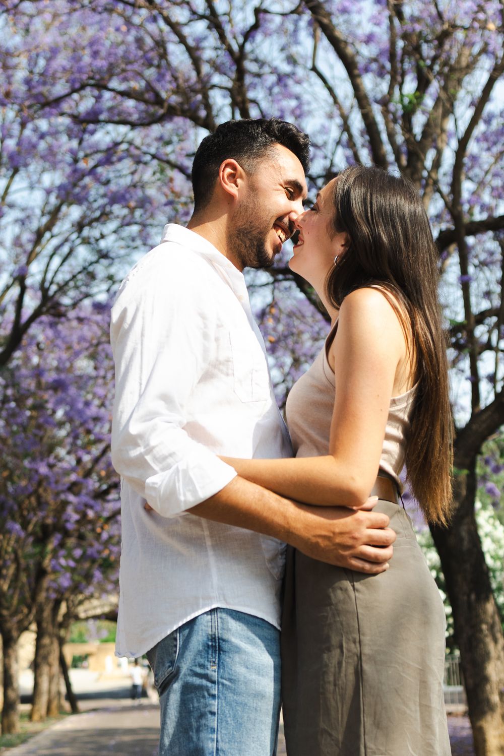 Couple sharing romantic kisses under blooming purple jacaranda trees on a tree-lined path.