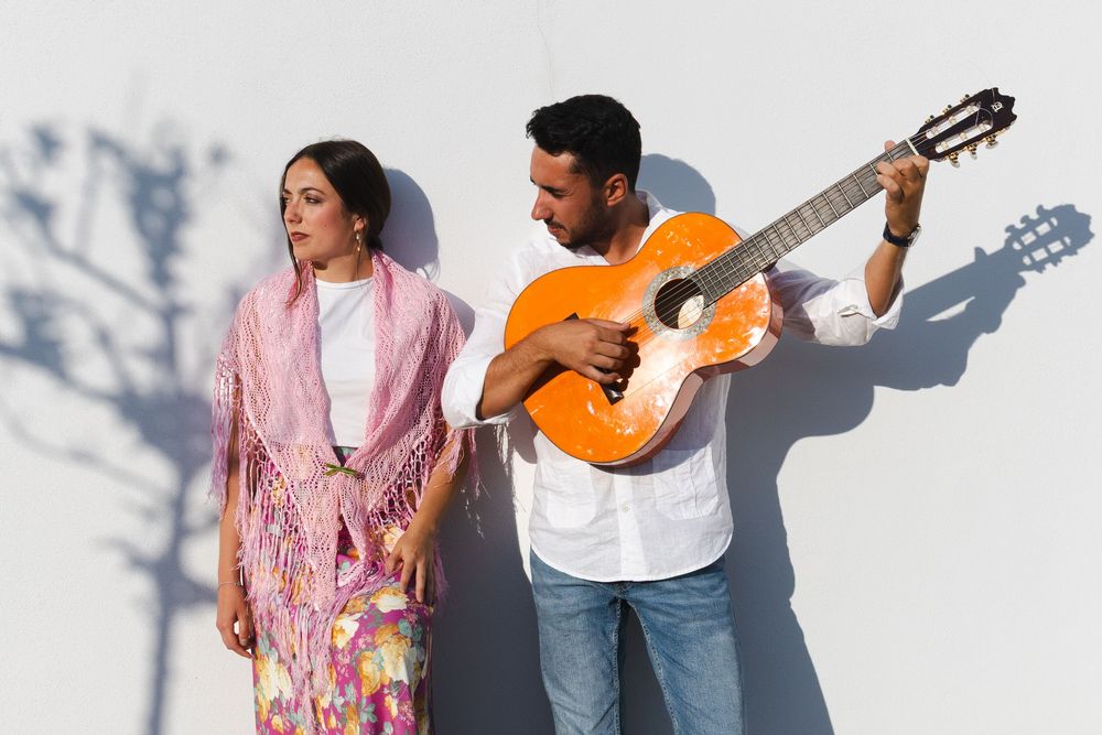A couple stands against a white wall while playing an acoustic guitar and dancing in sunlight.