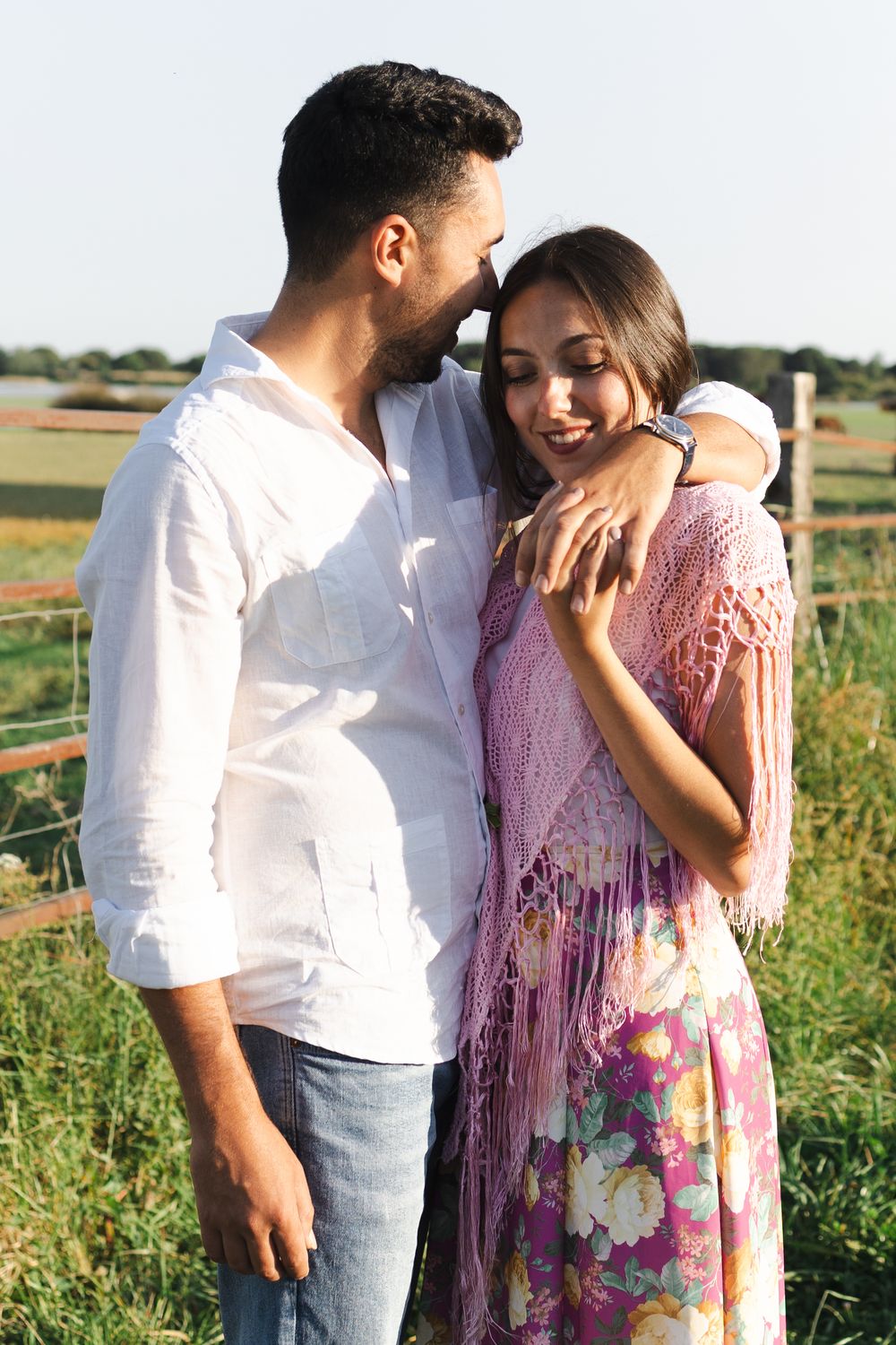 A couple shares intimate moments in a sunny field with tall grass and a rustic fence in the background.