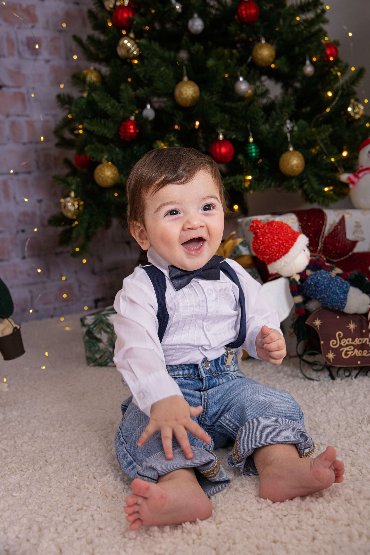 Baby in white shirt and denim overalls sits on carpet by Christmas tree with holiday decorations.
