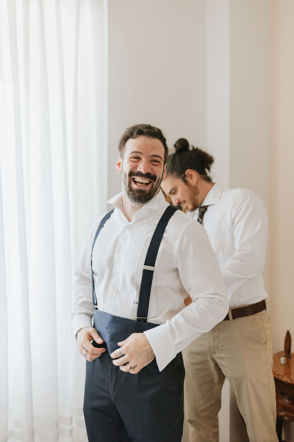 Two people in white dress shirts and suspenders share a candid laughing moment while getting ready.