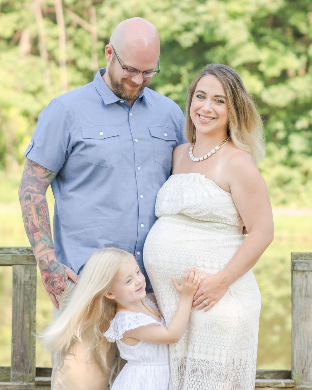A family maternity photo outdoors with a couple and young child posing together by a wooden fence.