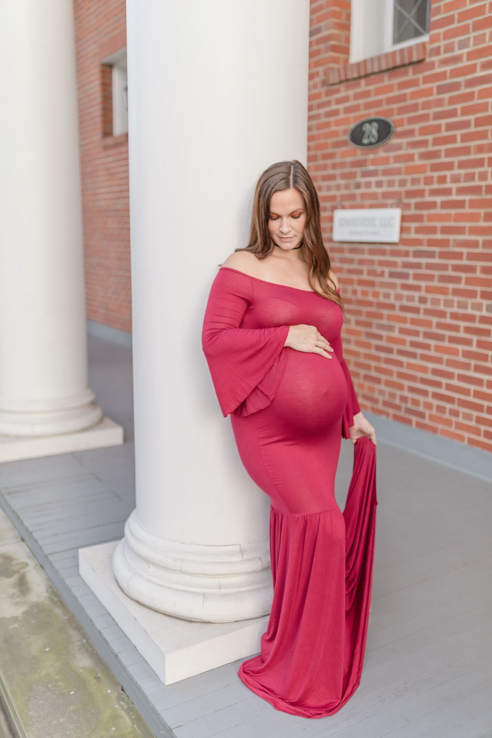 An expectant mother in a flowing red off-shoulder dress poses elegantly against a white column.