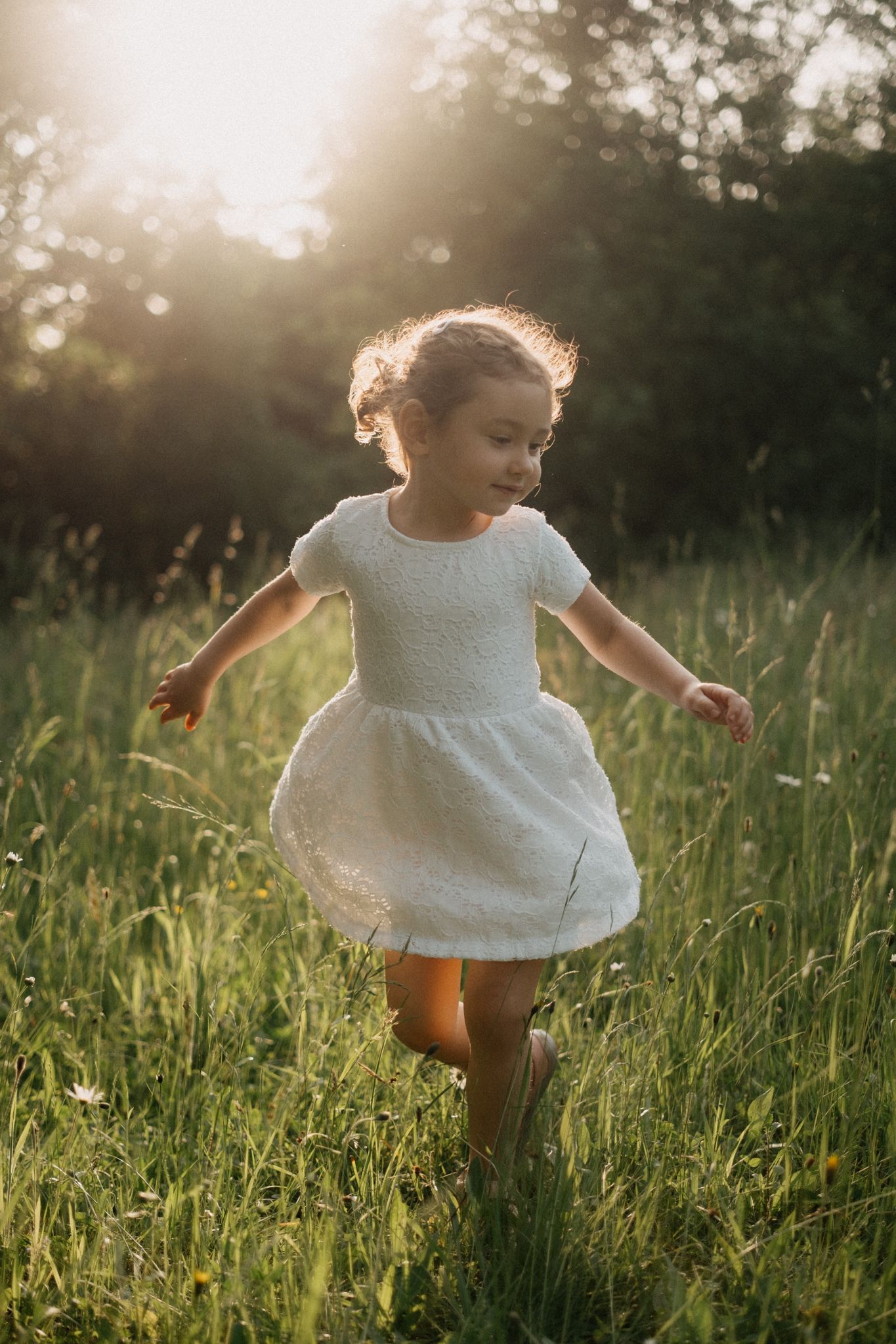 A small child in a white dress twirls through a sunlit meadow of tall grass during golden hour.