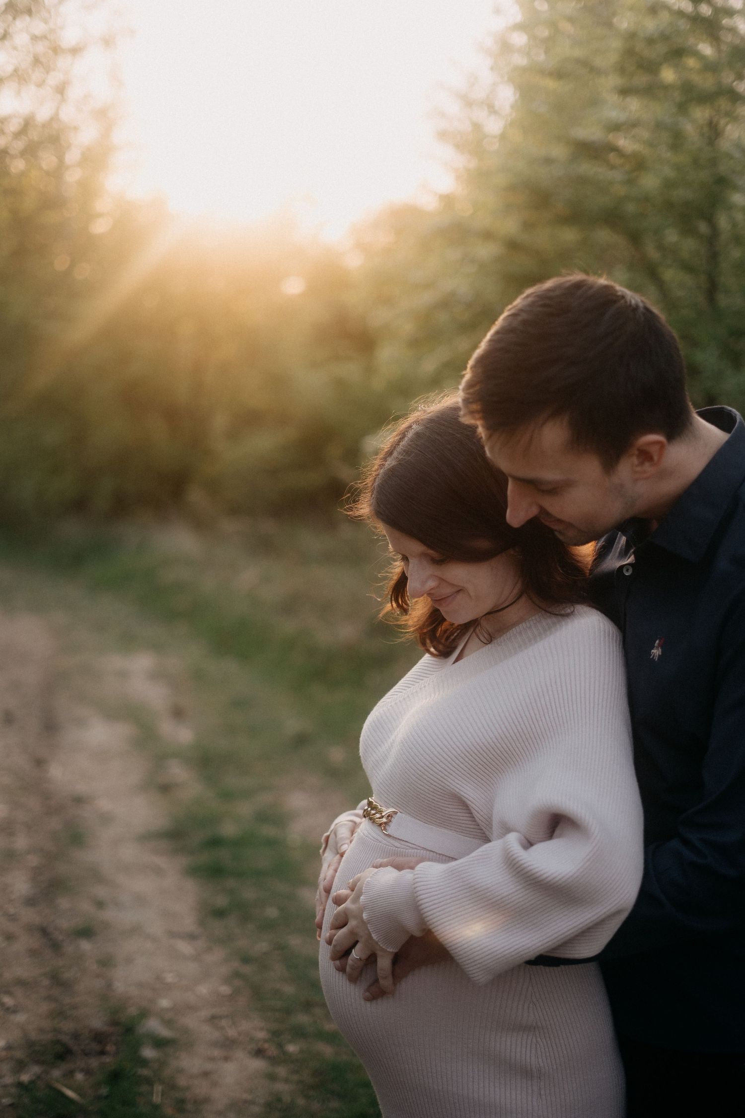 A couple embraces tenderly on a dirt path at sunset with golden light filtering through trees in the background.
