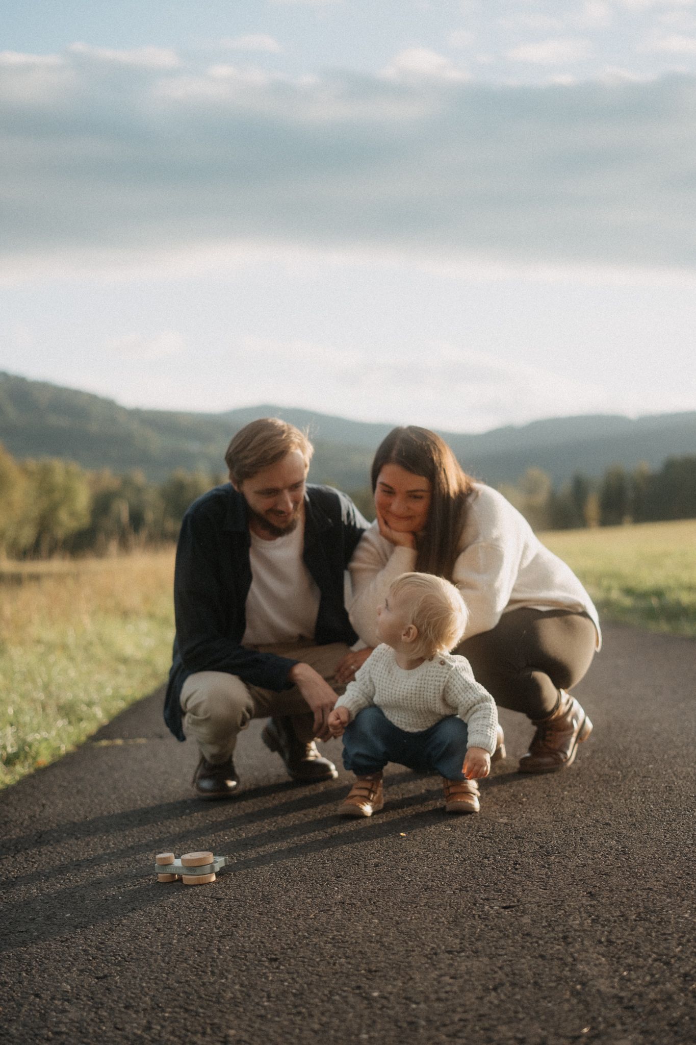 A couple crouches down together on a mountain road examining something small on the ground at sunset.