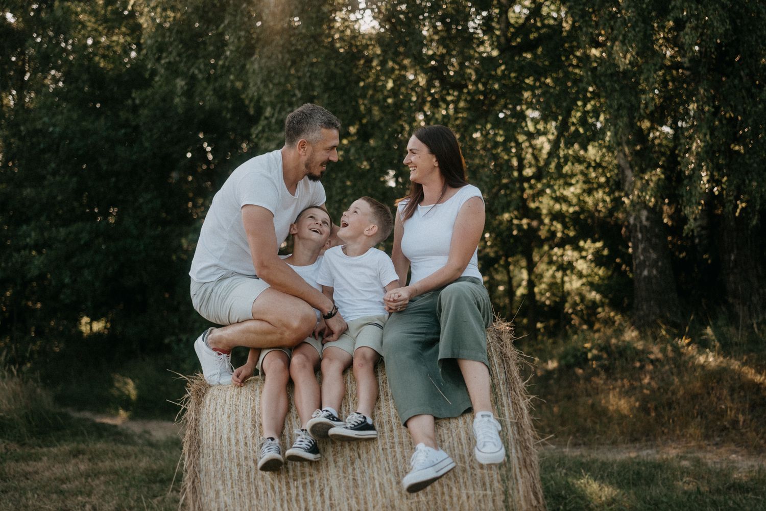 Family sitting together on a hay bale during golden hour in a rustic outdoor setting.