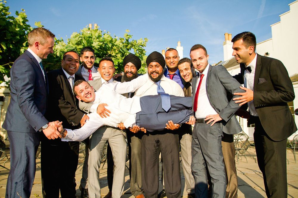 A joyful group of groomsmen in suits gather around and celebrate outdoors on a sunny day.