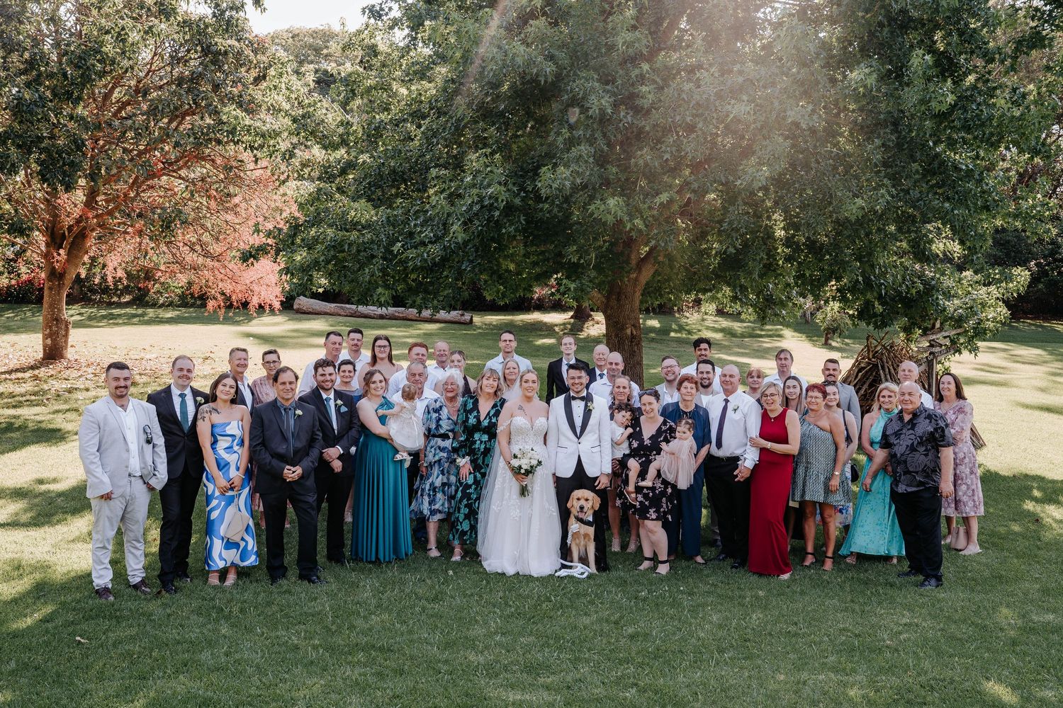 Wedding party and guests gather for group photos in a scenic outdoor garden setting on a sunny day.