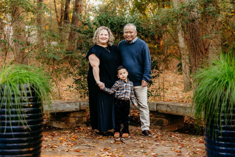A family of three poses together on a stone patio surrounded by autumn foliage and decorative grasses.