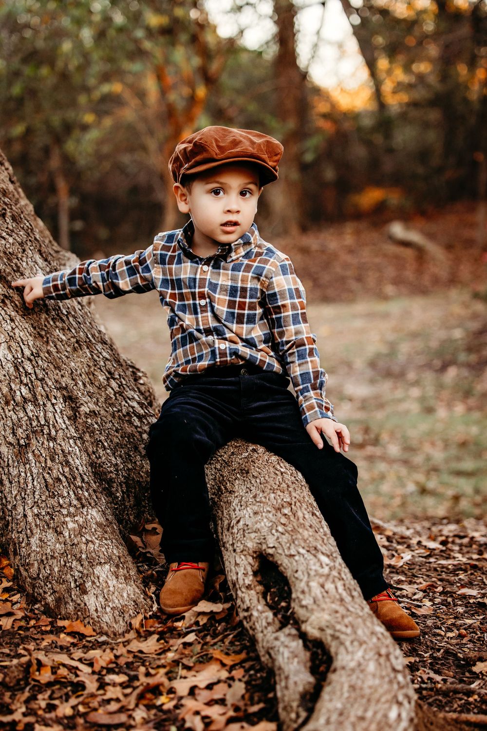 A child in a plaid shirt and overalls sits casually on a fallen tree trunk during autumn.