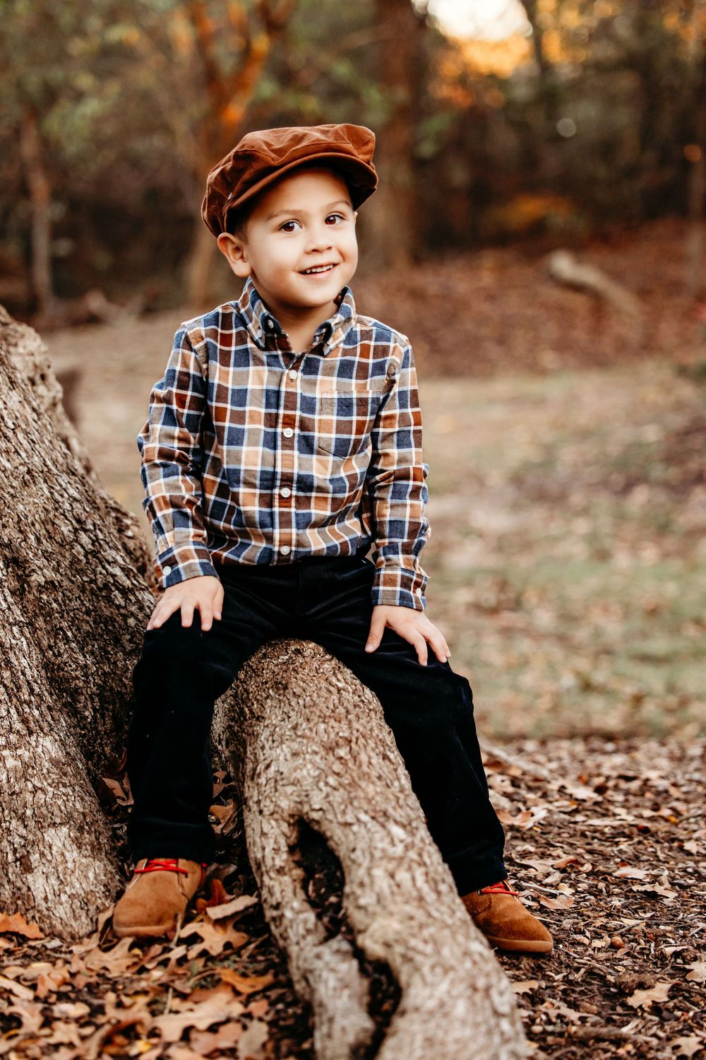 A child in a plaid shirt and baseball cap sits on a fallen tree trunk surrounded by autumn leaves.