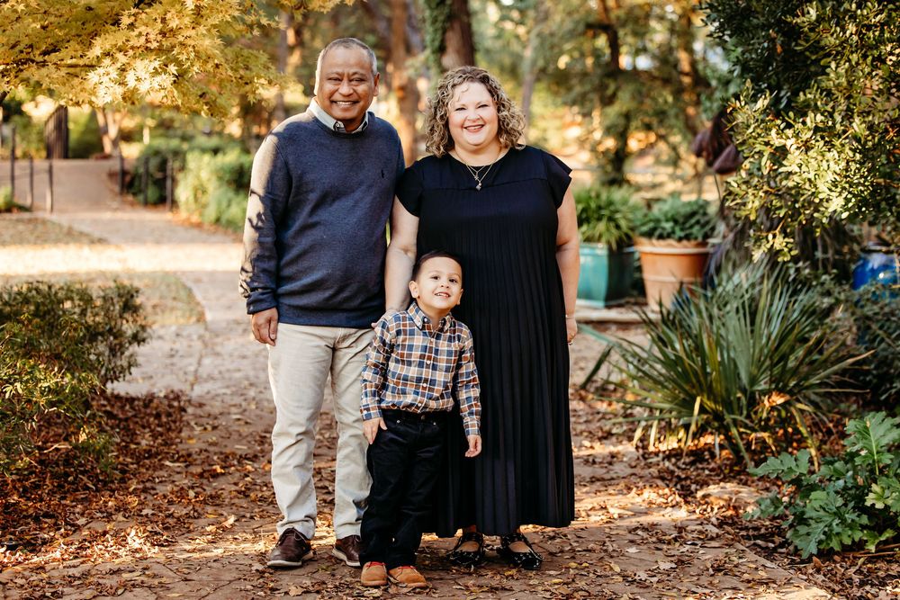 A family stands together on a garden path surrounded by autumn foliage and potted plants on a sunny day.