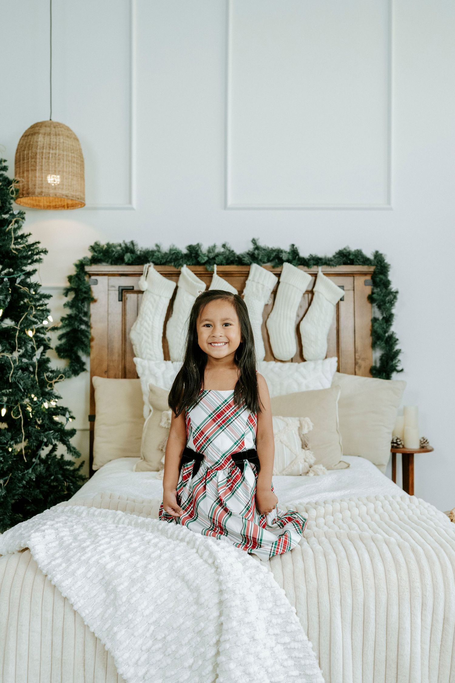 Child in plaid holiday dress sitting on white bed with Christmas stockings and greenery decorations in background.