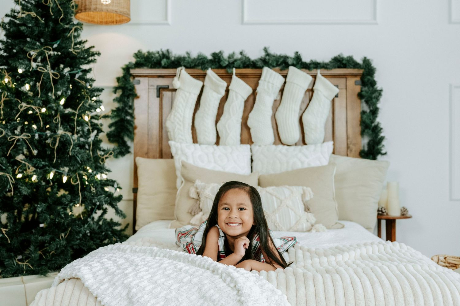 A cozy Christmas bedroom scene with a decorated tree, white bedding, and holiday stockings hanging on a wooden headboard.