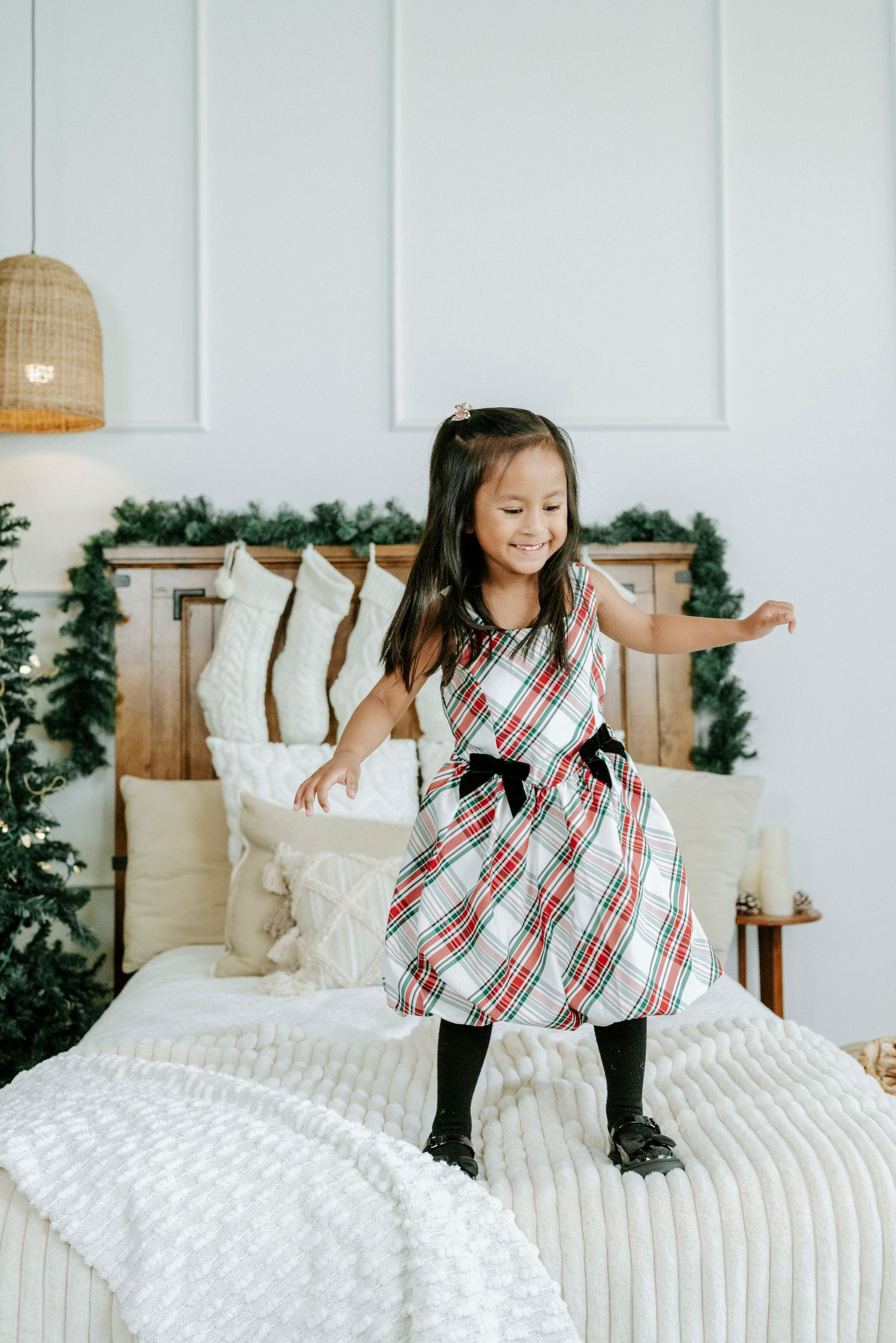 Child in plaid dress jumping on white bed with Christmas stockings and festive decor visible behind.