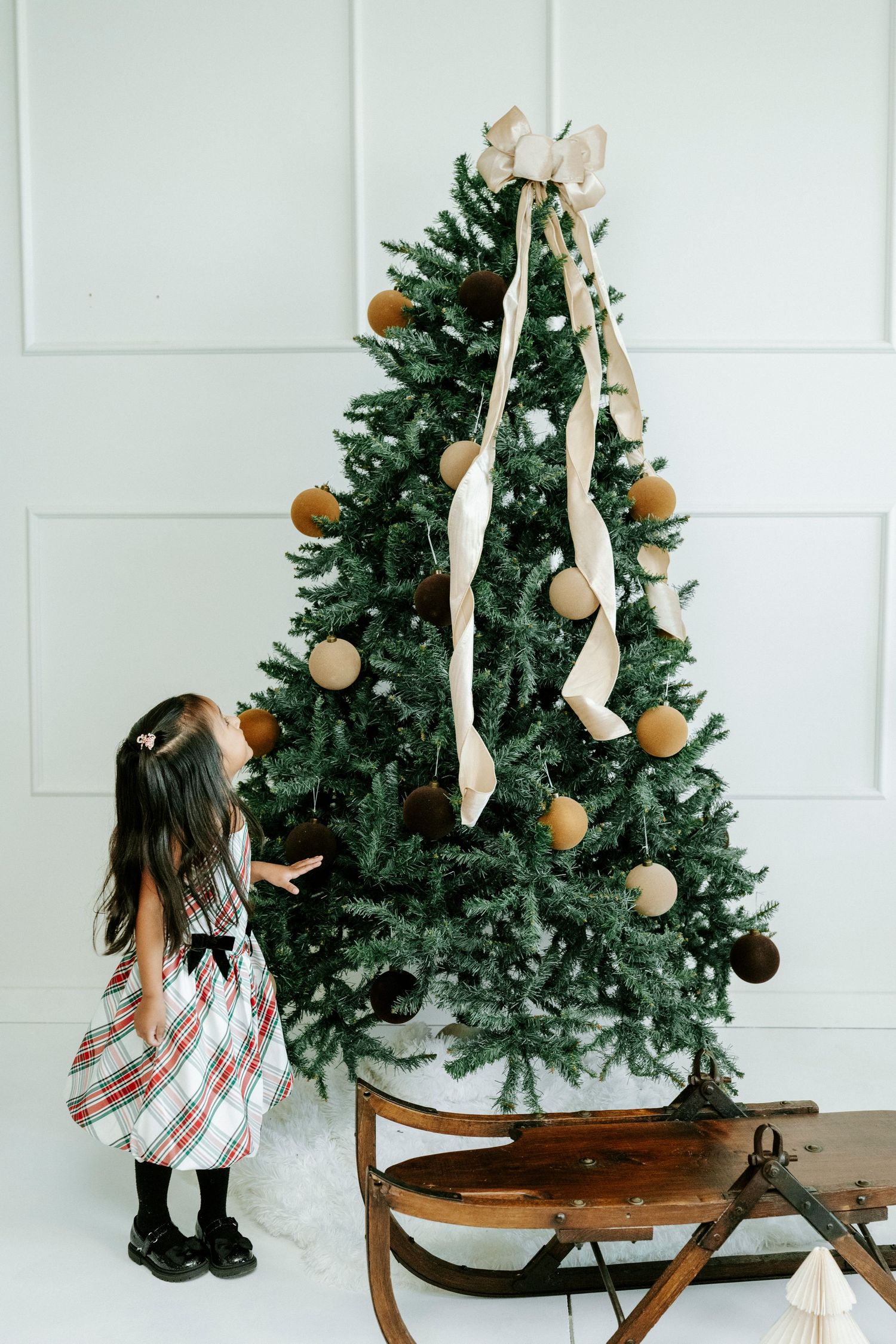 A child in a plaid dress decorates a Christmas tree placed on an antique wooden sled against a white wall.
