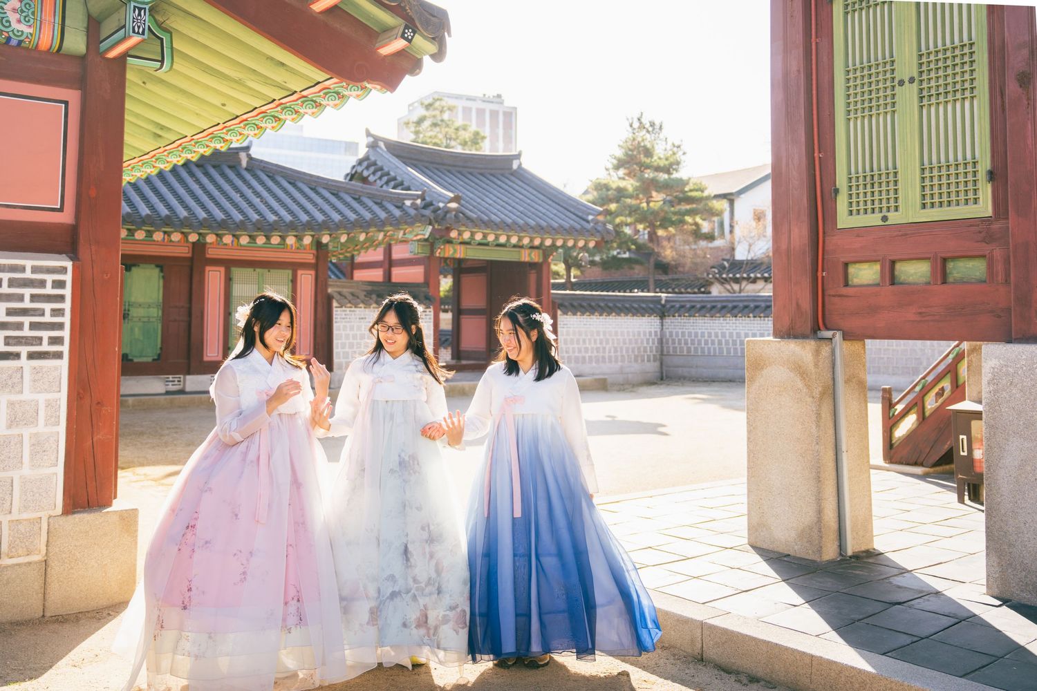 People wearing traditional Korean hanbok dresses walk through a historic palace courtyard on a sunny day.