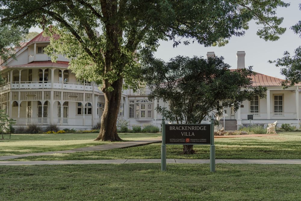 Historic white Victorian mansion with wrap-around porch surrounded by lush green lawn and mature trees.