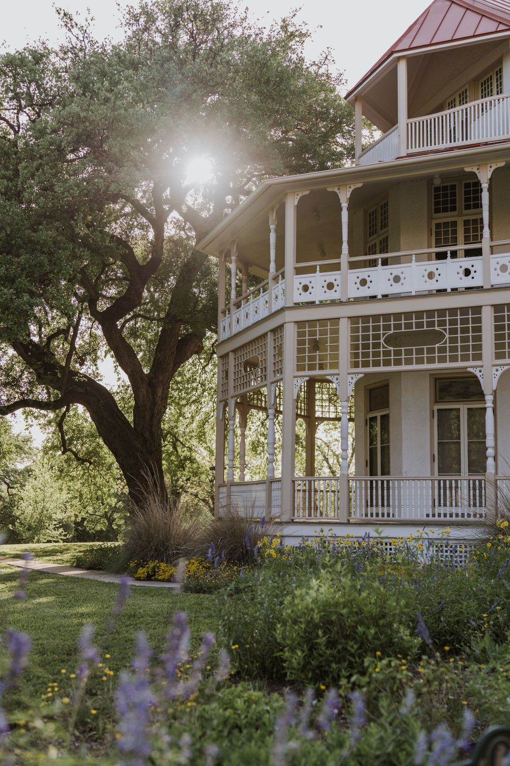 White colonial house with wrap-around porch and purple wildflowers blooming in the garden.