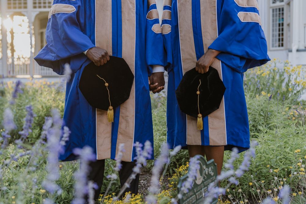 Row of graduates in bright blue academic regalia standing among purple flowers.