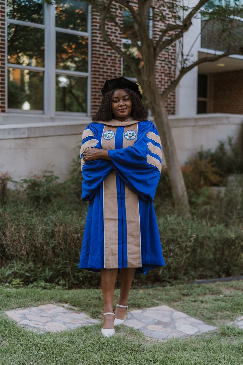 Graduate in blue and tan doctoral regalia standing on campus grounds.