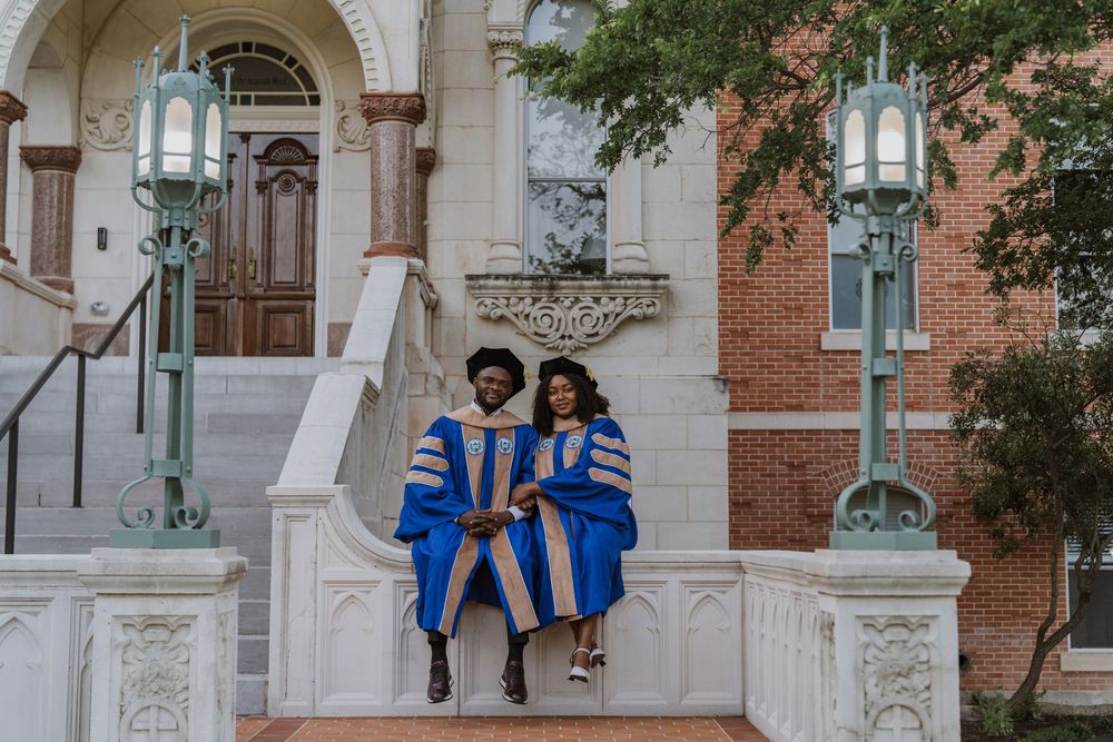 Two graduates in blue and gold academic regalia sitting on stone steps of university building.