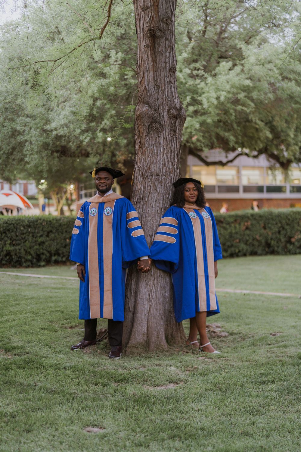 Two graduates in blue doctoral robes posing against a tree trunk on campus lawn.