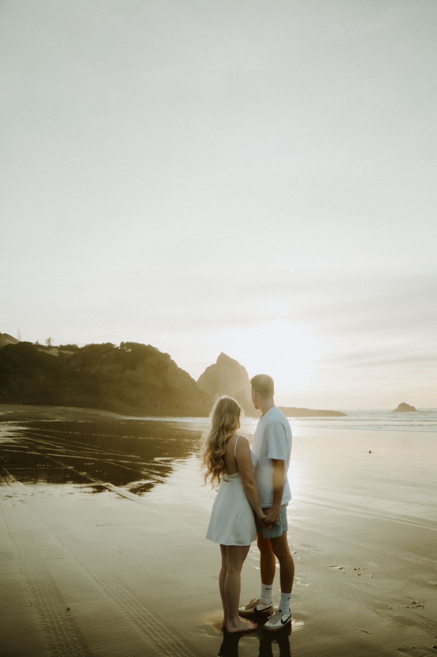Couple walks hand in hand along a serene beach during golden hour with cliffs in the background.